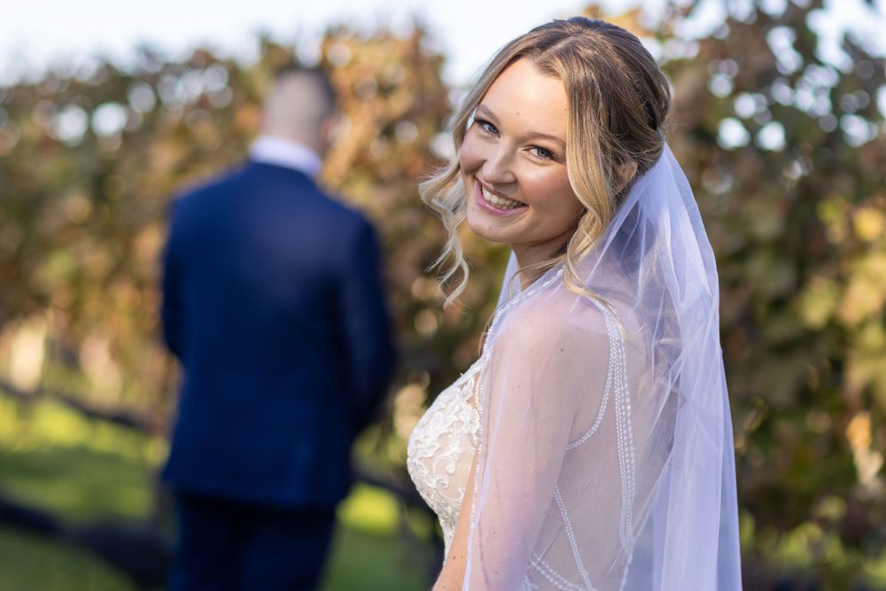 Smiling bride in lace gown and veil looks back at camera with groom blurred in background.