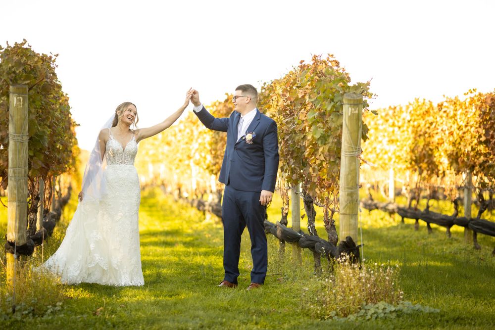 Bride and groom holding hands in golden autumn vineyard, bride in lace gown, groom in navy suit.
