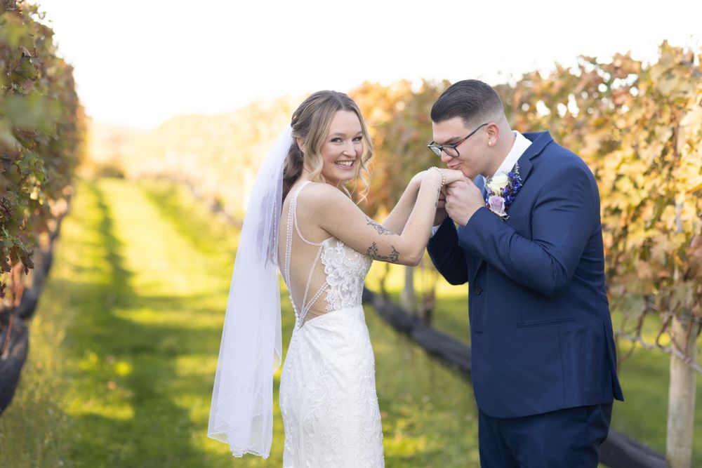 Groom in navy suit kissing brides hand in sunlit vineyard as bride laughs in lace wedding gown.