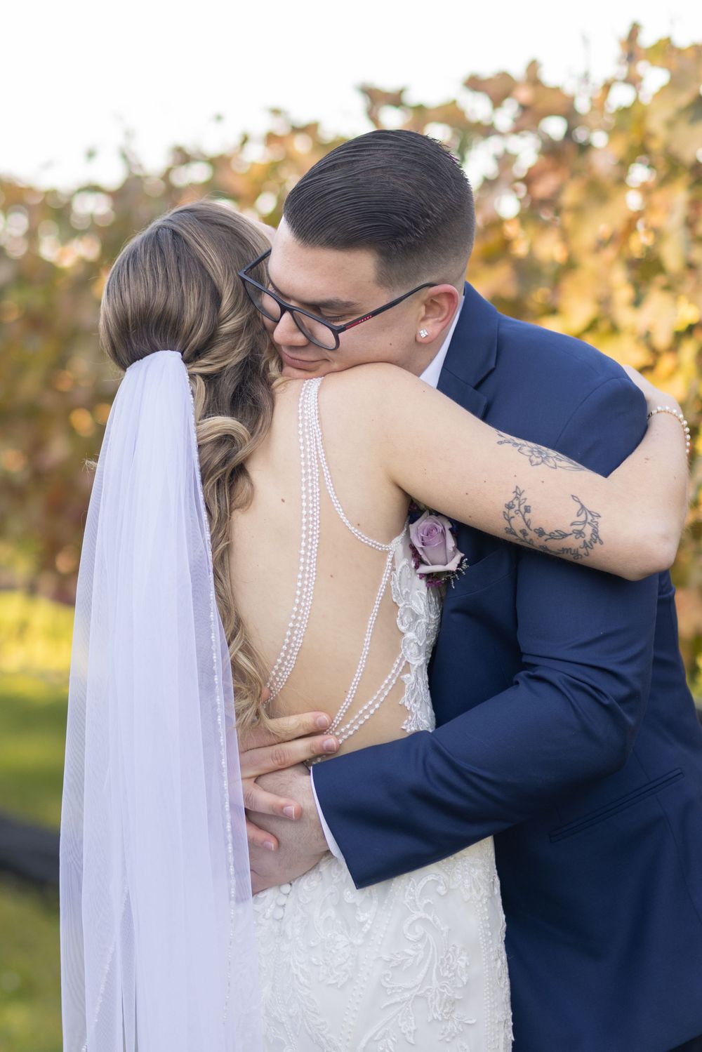 Groom in navy suit embracing bride showing pearl-detailed open back lace wedding gown in vineyard.