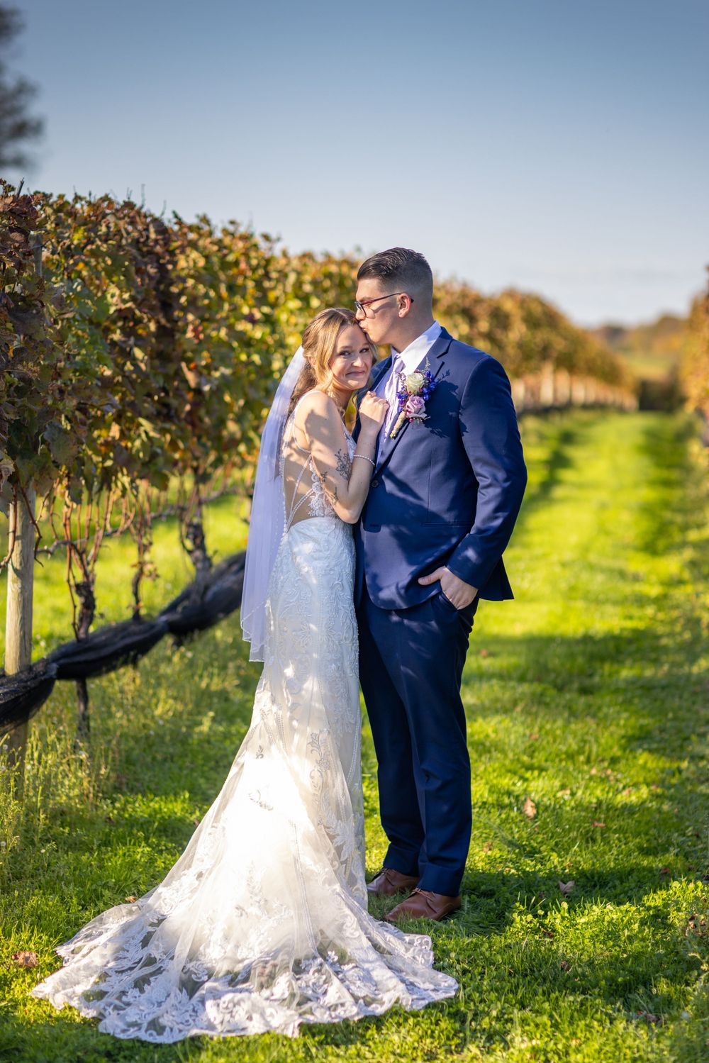 Groom in navy suit kissing brides temple as she leans into him in sunny autumn vineyard setting.
