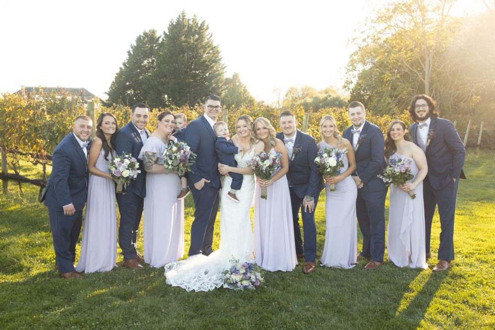 Wedding party poses together on green lawn at golden hour, bridesmaids in lavender gowns and groomsmen in navy suits.