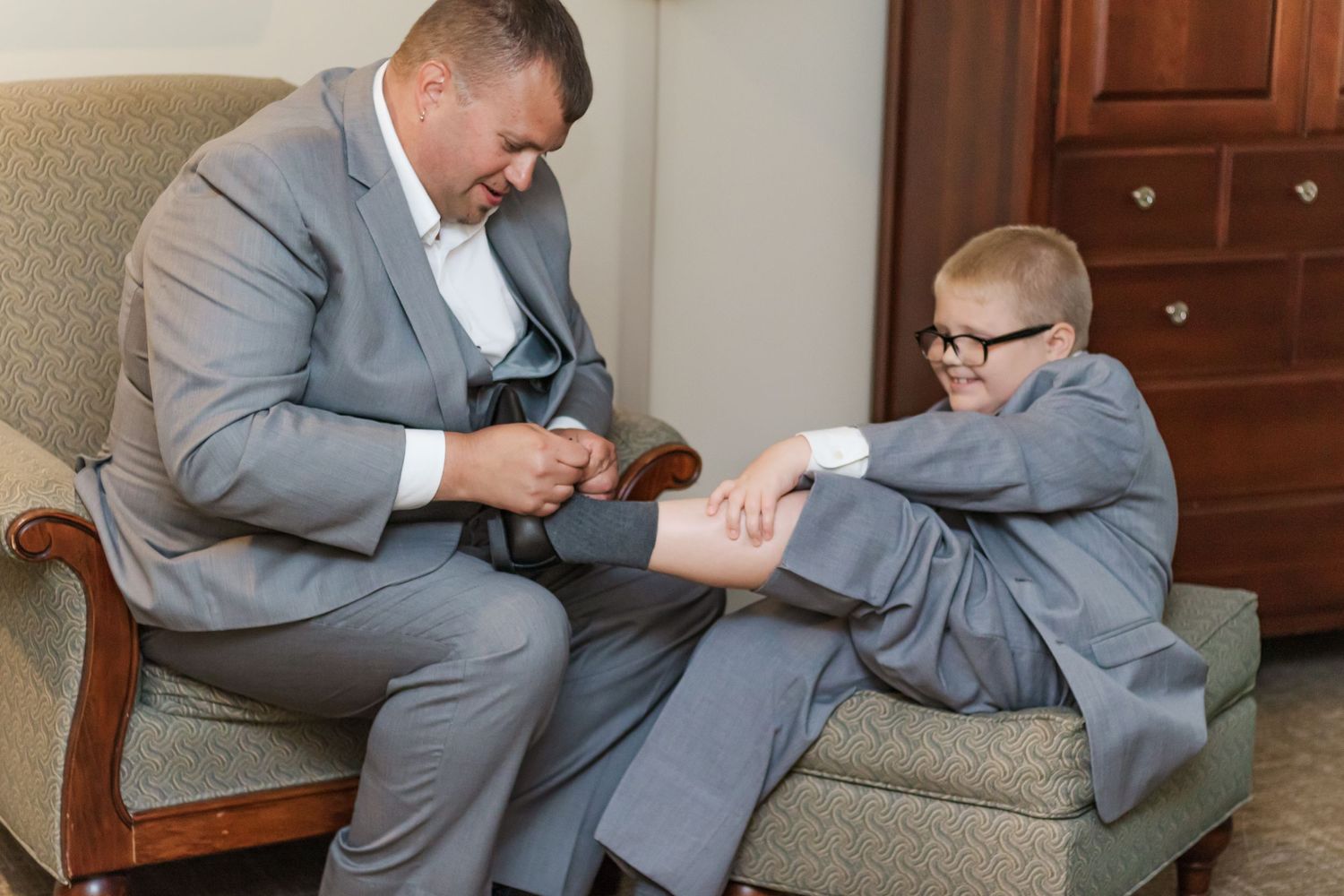 Two people in gray suits sharing a light moment while getting ready.