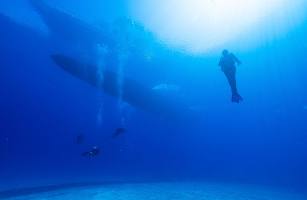 Scuba divers swimming beneath large boat silhouettes in blue ocean waters.
