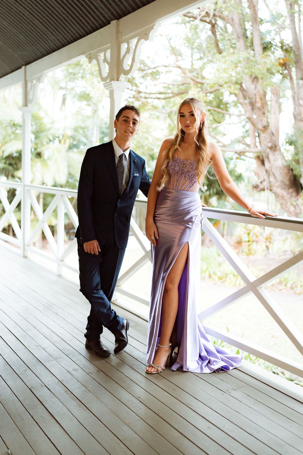 A couple in formal attire poses on a wooden veranda, wearing a navy suit and lavender gown with floral bodice.