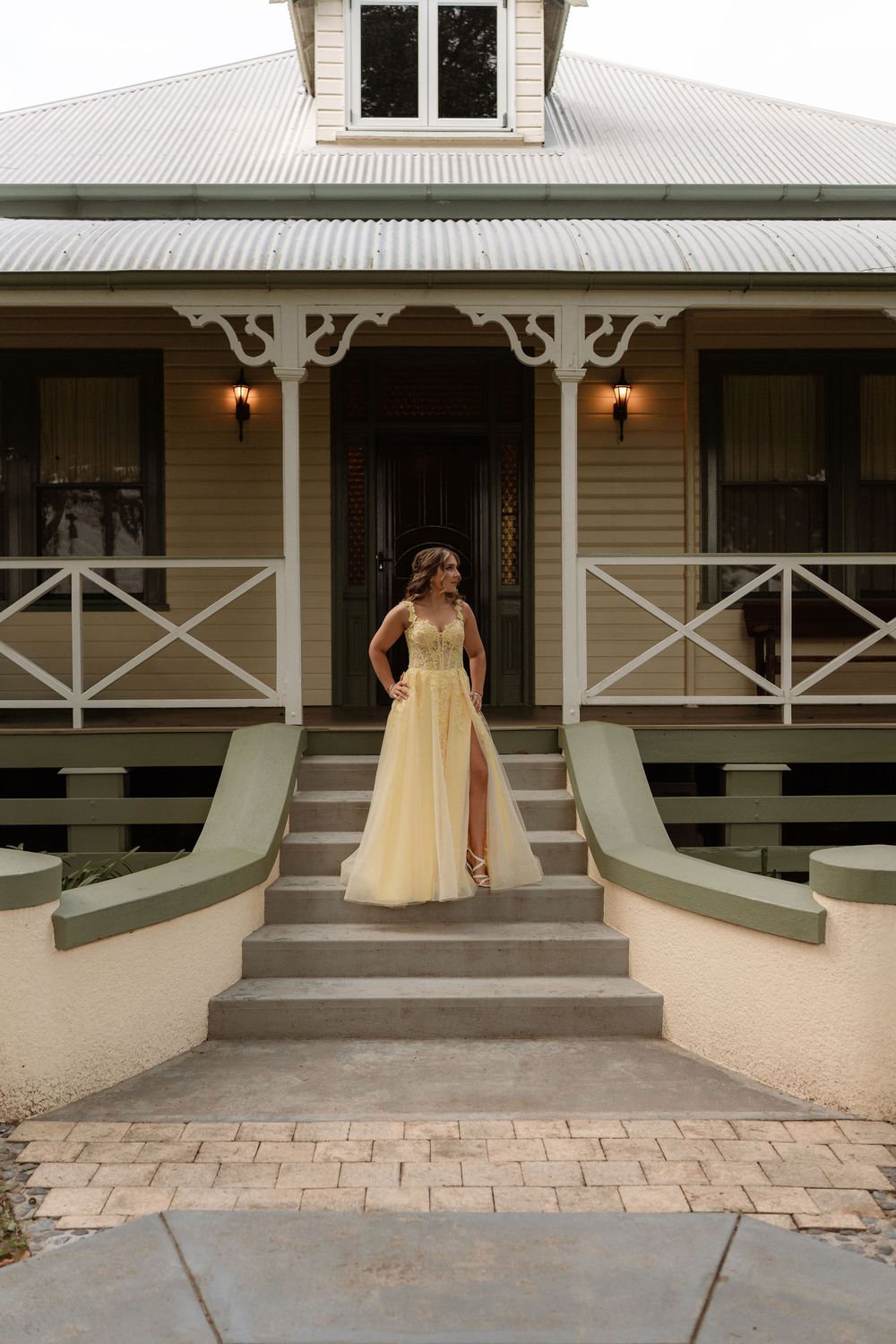 Woman in yellow ball gown posing on steps of charming heritage home with decorative white verandah.