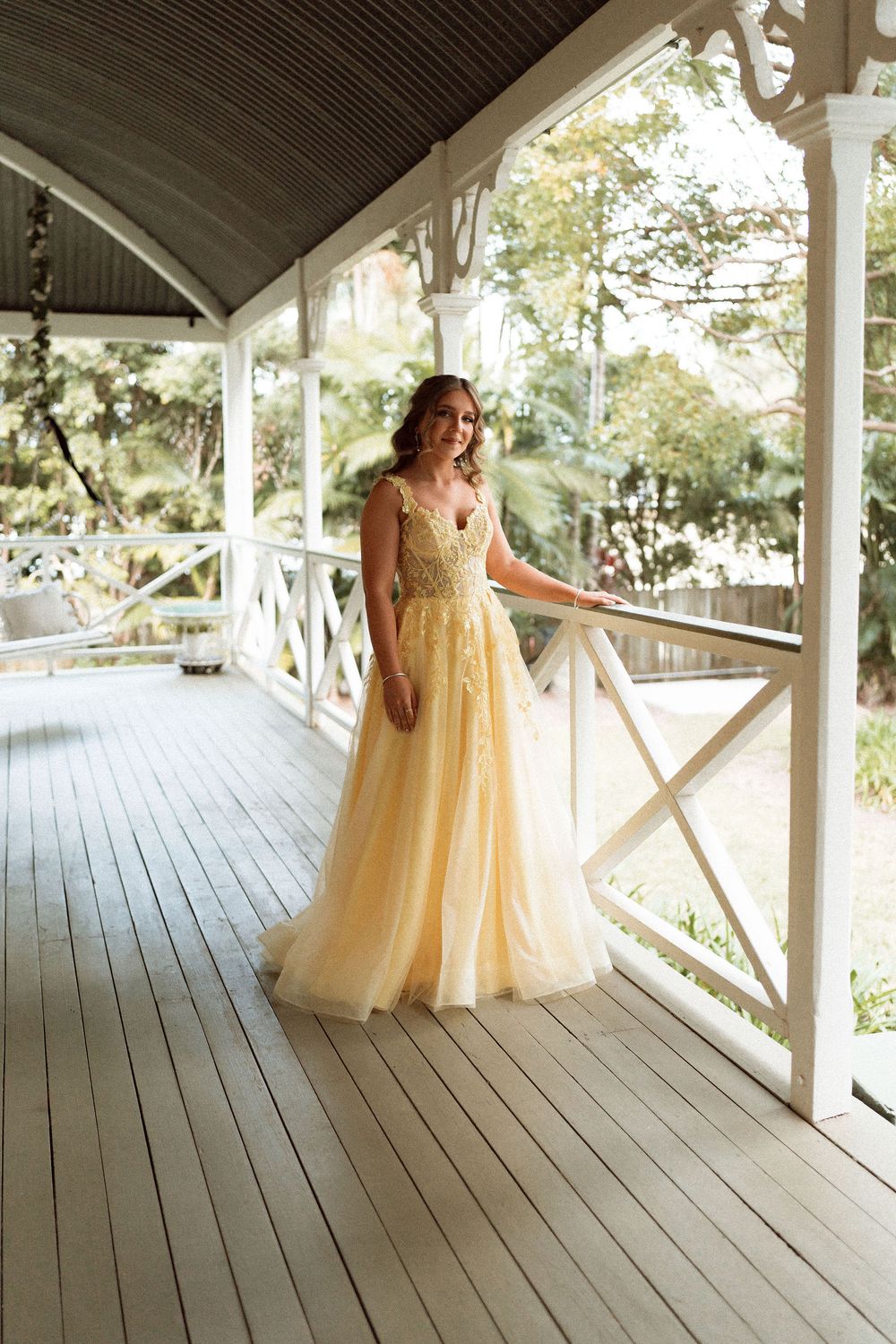 Woman in yellow tulle ball gown standing on white wooden porch surrounded by lush greenery.