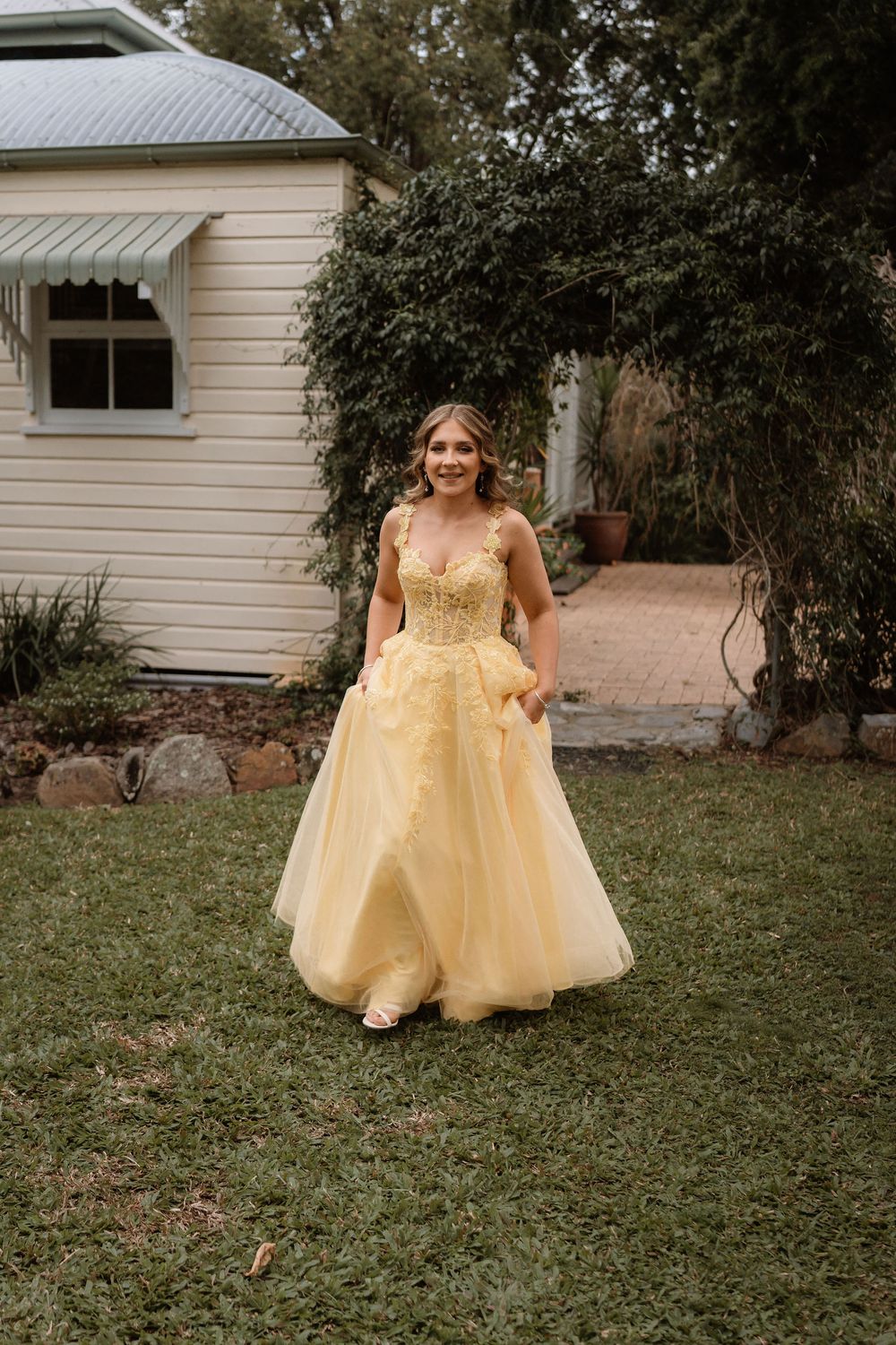Young woman in a yellow ball gown posing outdoors on green lawn beside a white house.