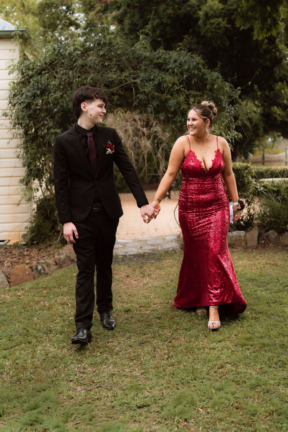 Couple in formal attire walking hand in hand, man in black suit, woman in red sequin gown, outdoor garden setting.