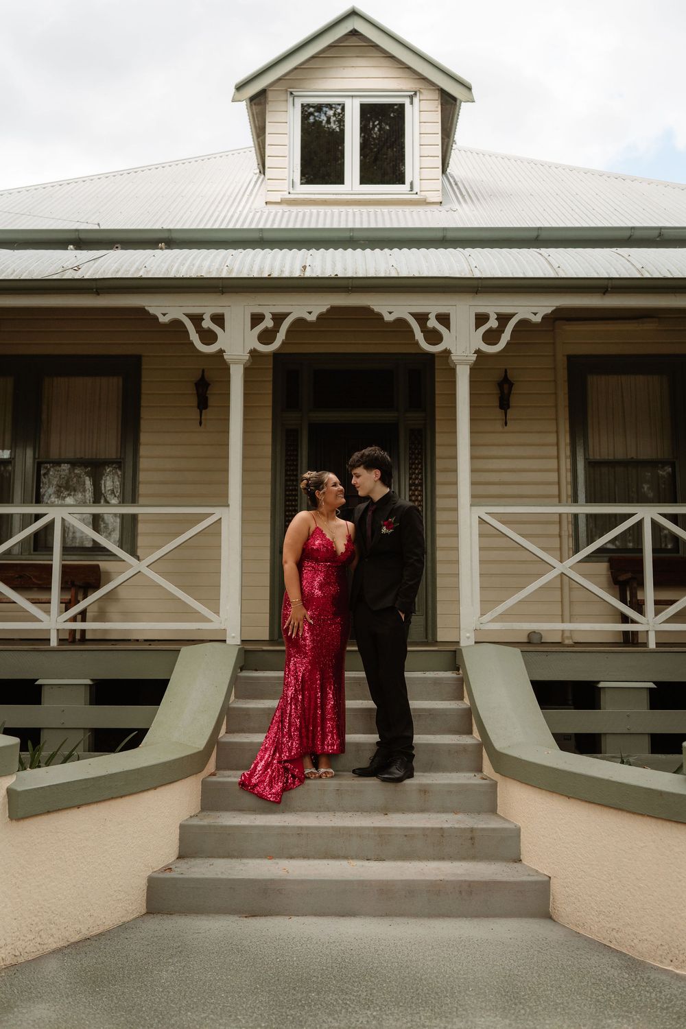 A couple in formal attire pose on steps of a charming Queensland heritage home, woman in red sequin gown, man in black suit.
