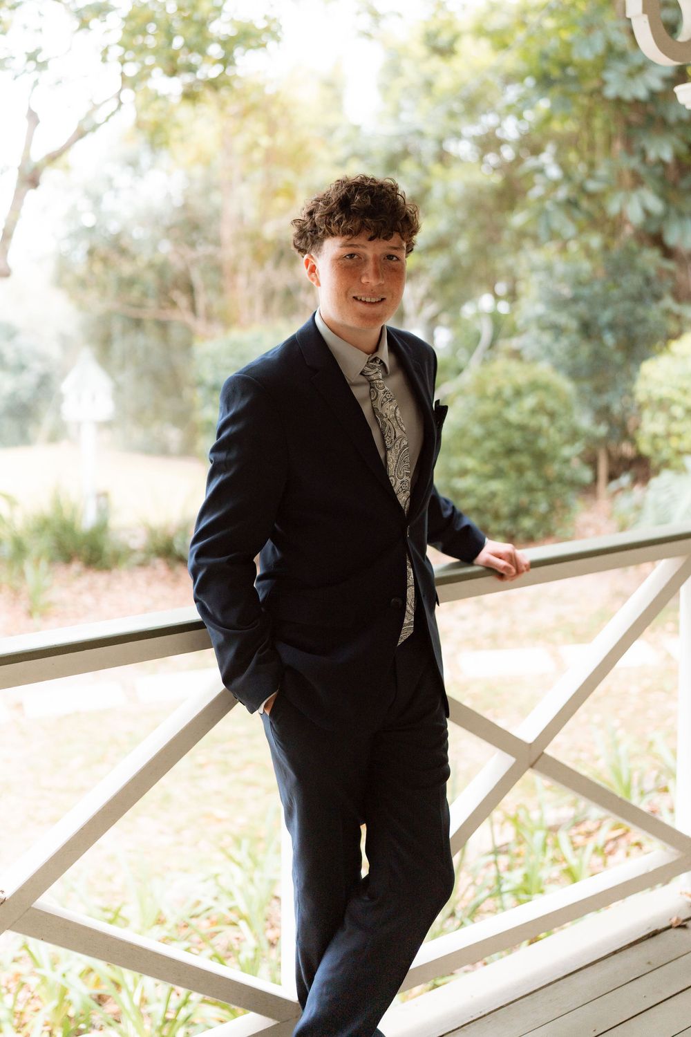 Young man in navy suit and patterned tie smiling on a wooden bridge surrounded by lush greenery.