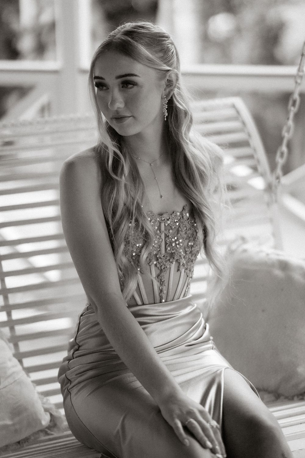 Black and white photo of a young blonde woman in a beaded gown sitting elegantly on a white porch swing.