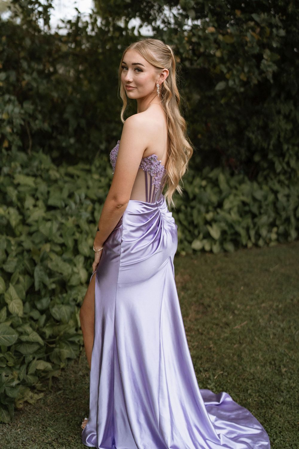 Young woman in elegant lavender satin prom gown with floral corset bodice and high slit, posing outdoors.