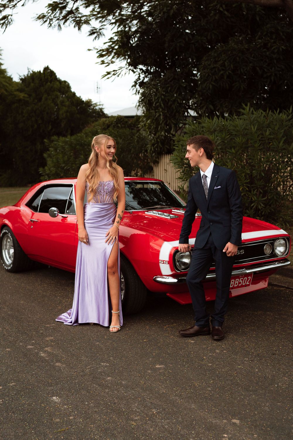 Couple in formal wear posing by a classic red Chevrolet Camaro, woman in lavender gown and man in navy suit.