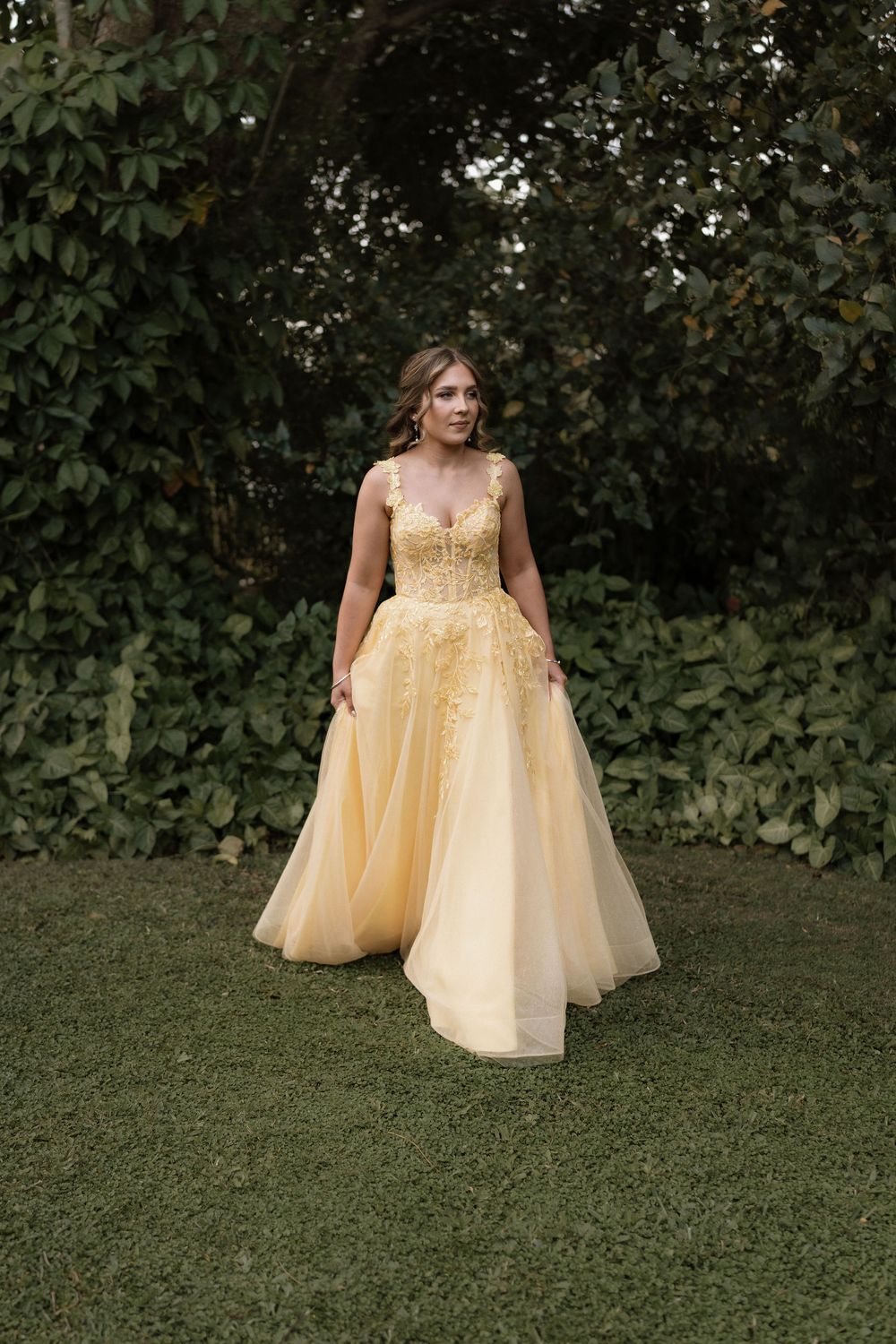 Woman in elegant yellow ball gown with floral applique bodice standing on grass before lush green foliage.