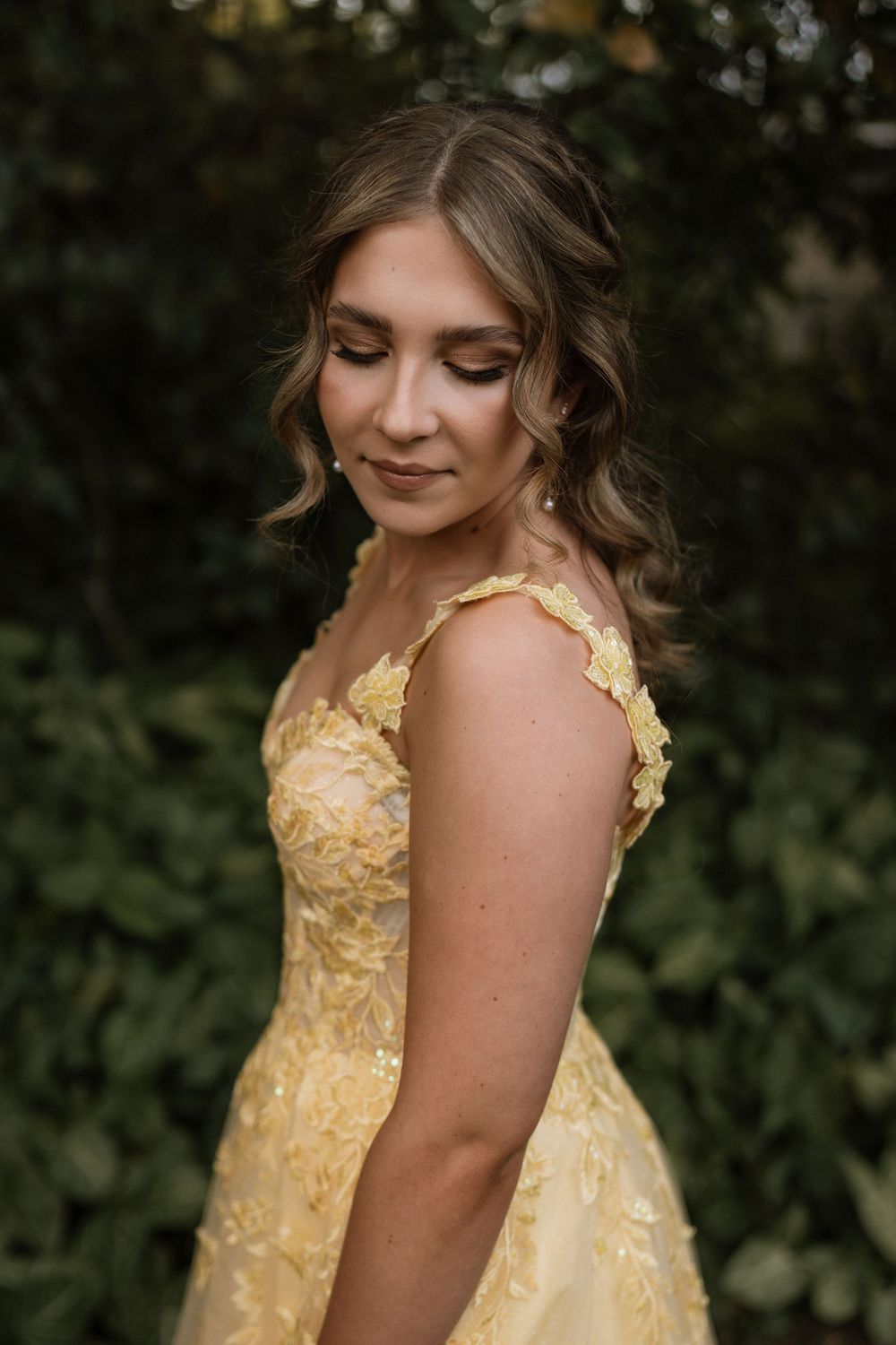 Young woman in elegant yellow floral gown posing outdoors with soft curled hair and natural makeup.