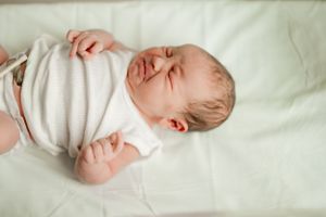 Newborn baby in white onesie crying while lying on a soft white surface in a hospital nursery.