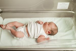 Newborn baby in white outfit lying in a hospital bassinet with arms and legs stretched out.