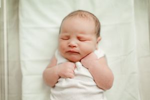 Sleeping newborn baby in a white onesie with hands tucked under chin lying in a hospital crib.