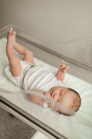 Newborn baby in white onesie lying in a hospital bassinet with legs and arms raised upward.