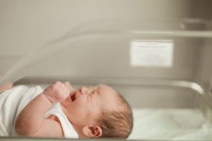 Newborn baby in white clothing lying in a hospital bassinet with mouth slightly open.