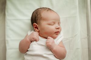 Peaceful newborn baby sleeping on white blanket wearing white onesie with tiny fists raised.
