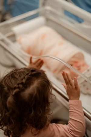 Young curly-haired girl in pink shirt looking at newborn baby in hospital bassinet.