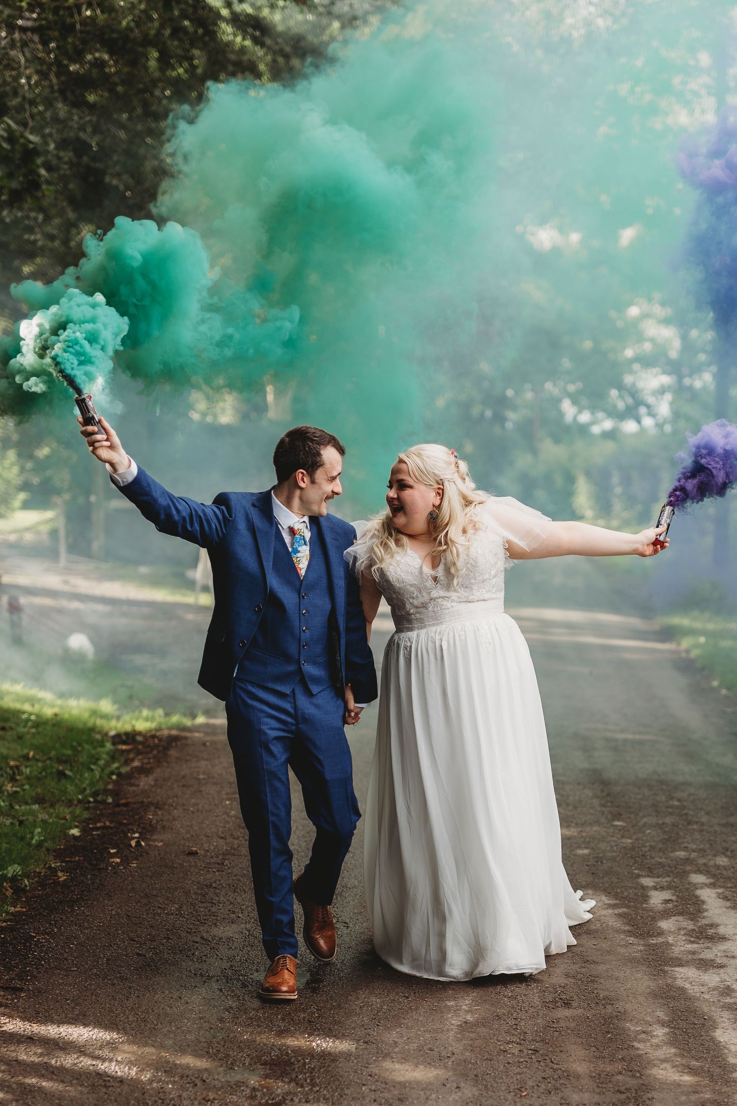 Newlyweds holding colored smoke bombs creating teal clouds during outdoor wedding photos.