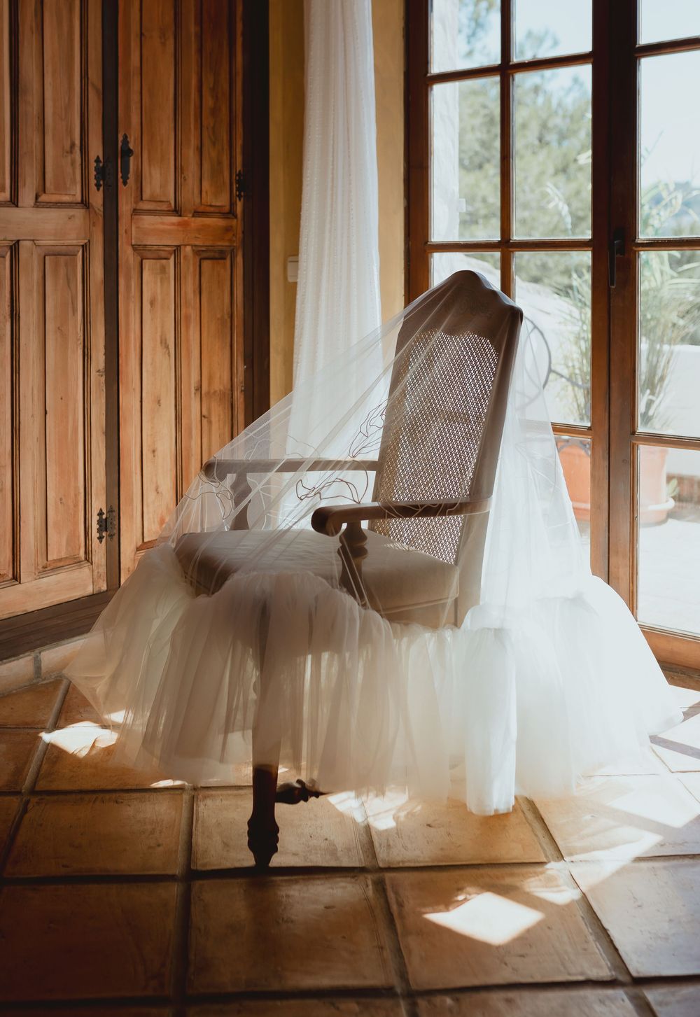 A flowing white wedding dress hangs elegantly in front of wooden doors and white curtains in natural light.