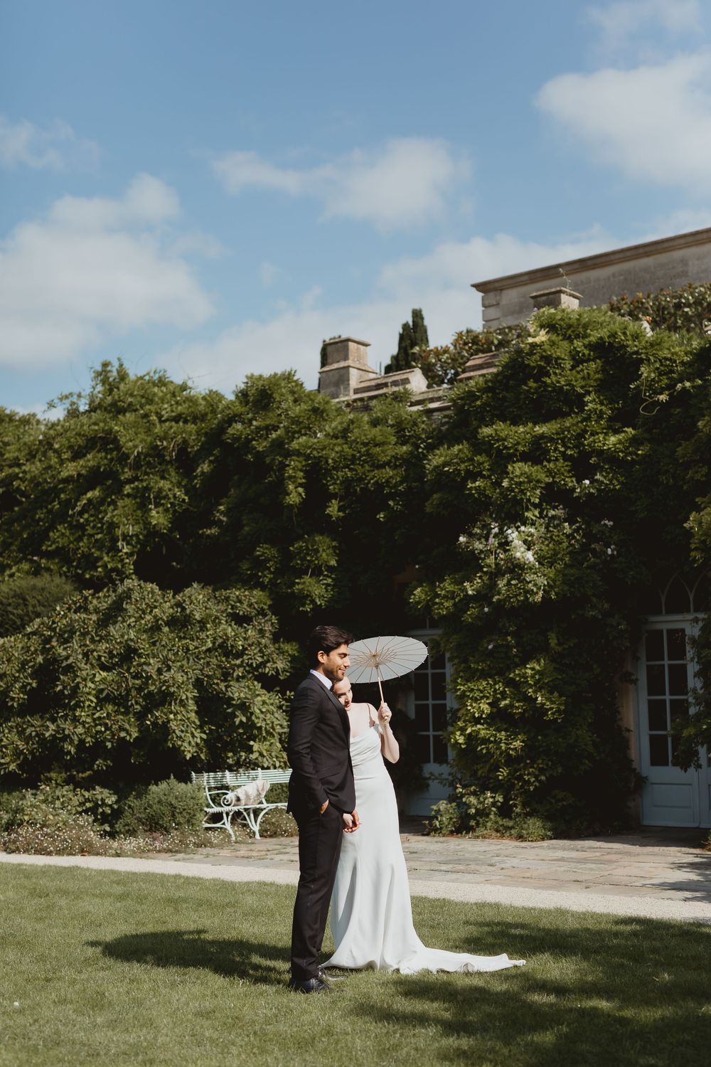 A couple stands together on a sunny day surrounded by lush greenery and architecture for their outdoor wedding.