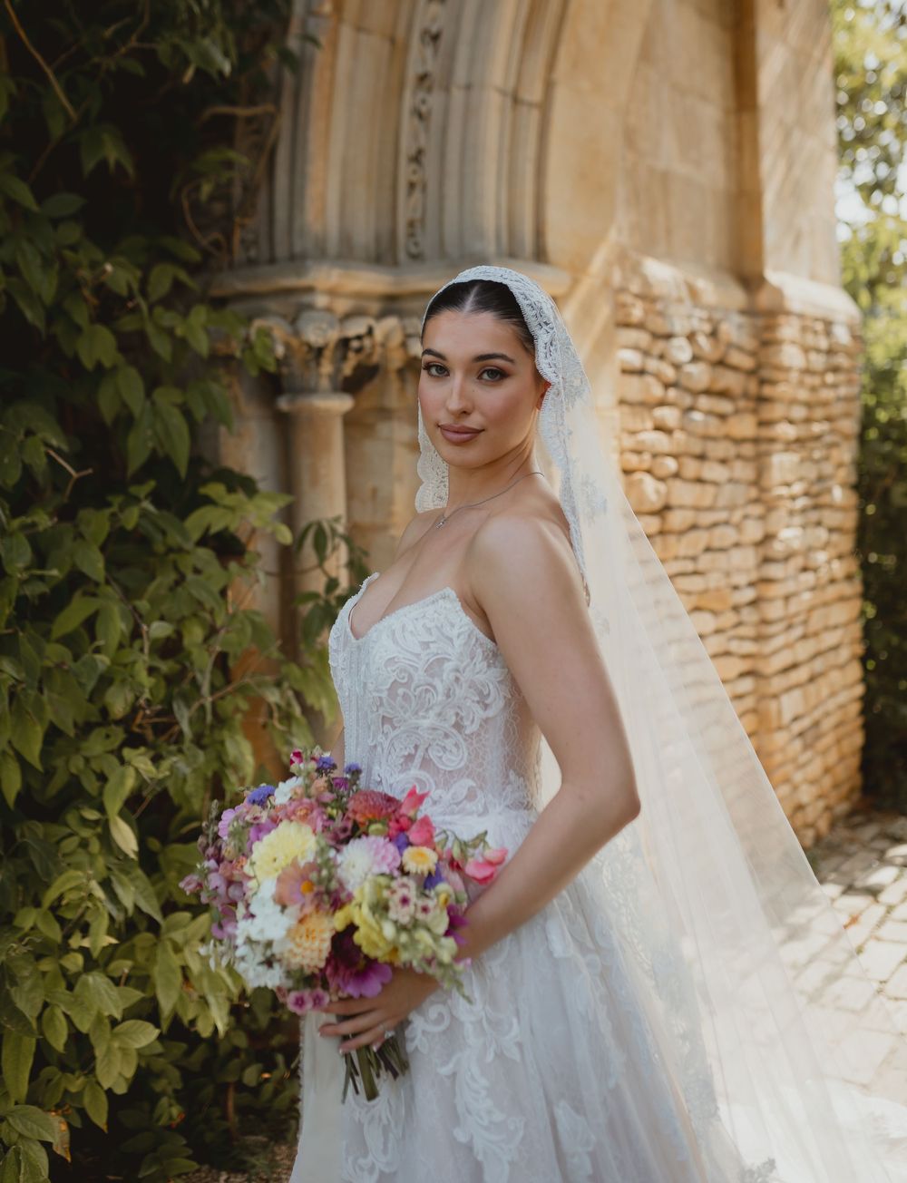 A bride in a white lace wedding gown holds a colorful bouquet of flowers against a rustic stone wall backdrop.