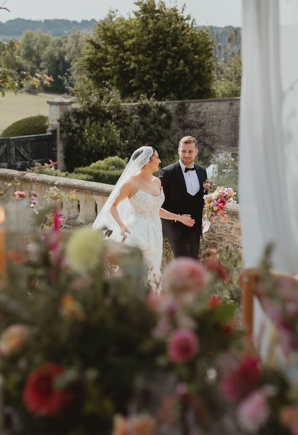 Two people share a romantic wedding moment in an outdoor garden setting framed by white columns and pink flowers.