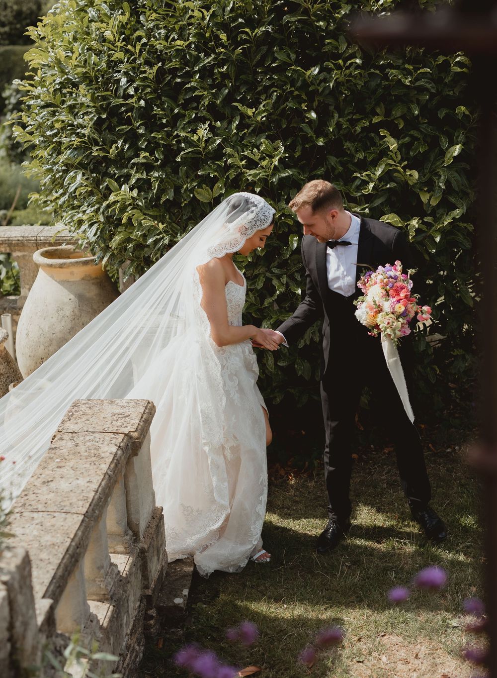 A bride in a flowing white dress explores a garden setting with stone walls and greenery.