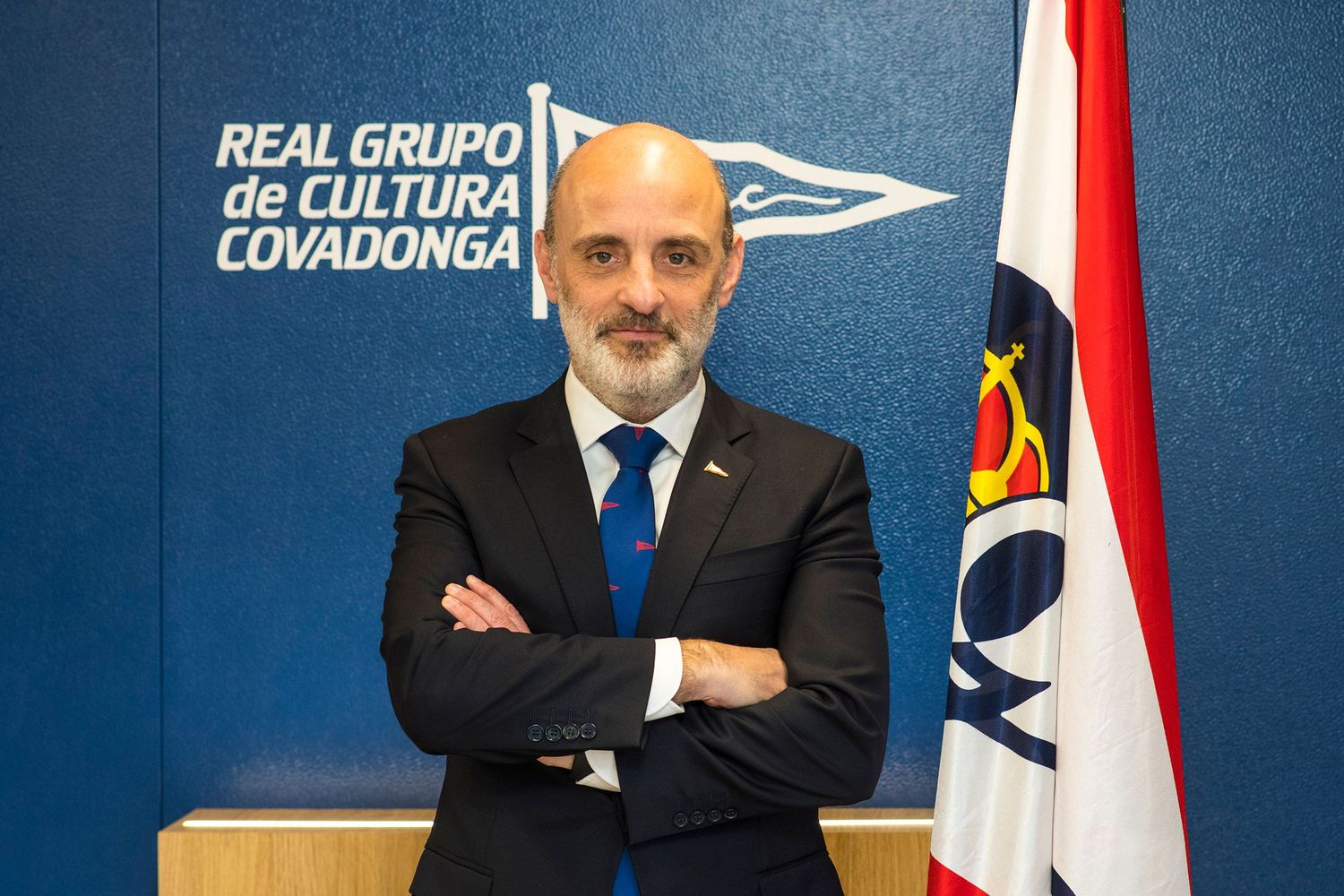 Un hombre con traje y corbata se encuentra con los brazos cruzados frente a un logotipo corporativo y una bandera.