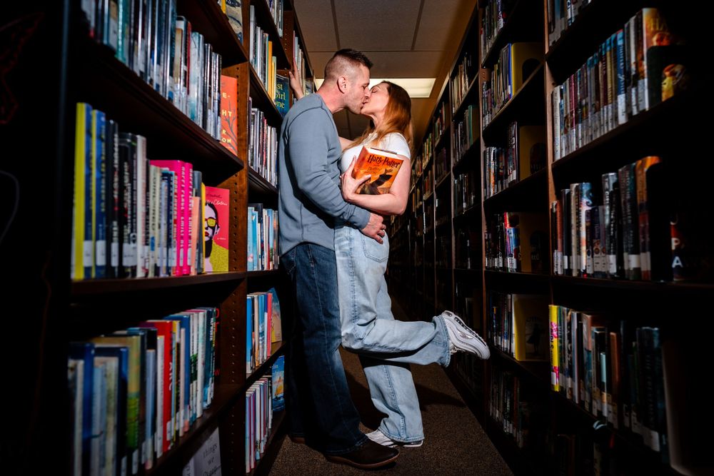 Silhouetted figures embracing in a dimly lit library aisle surrounded by books.