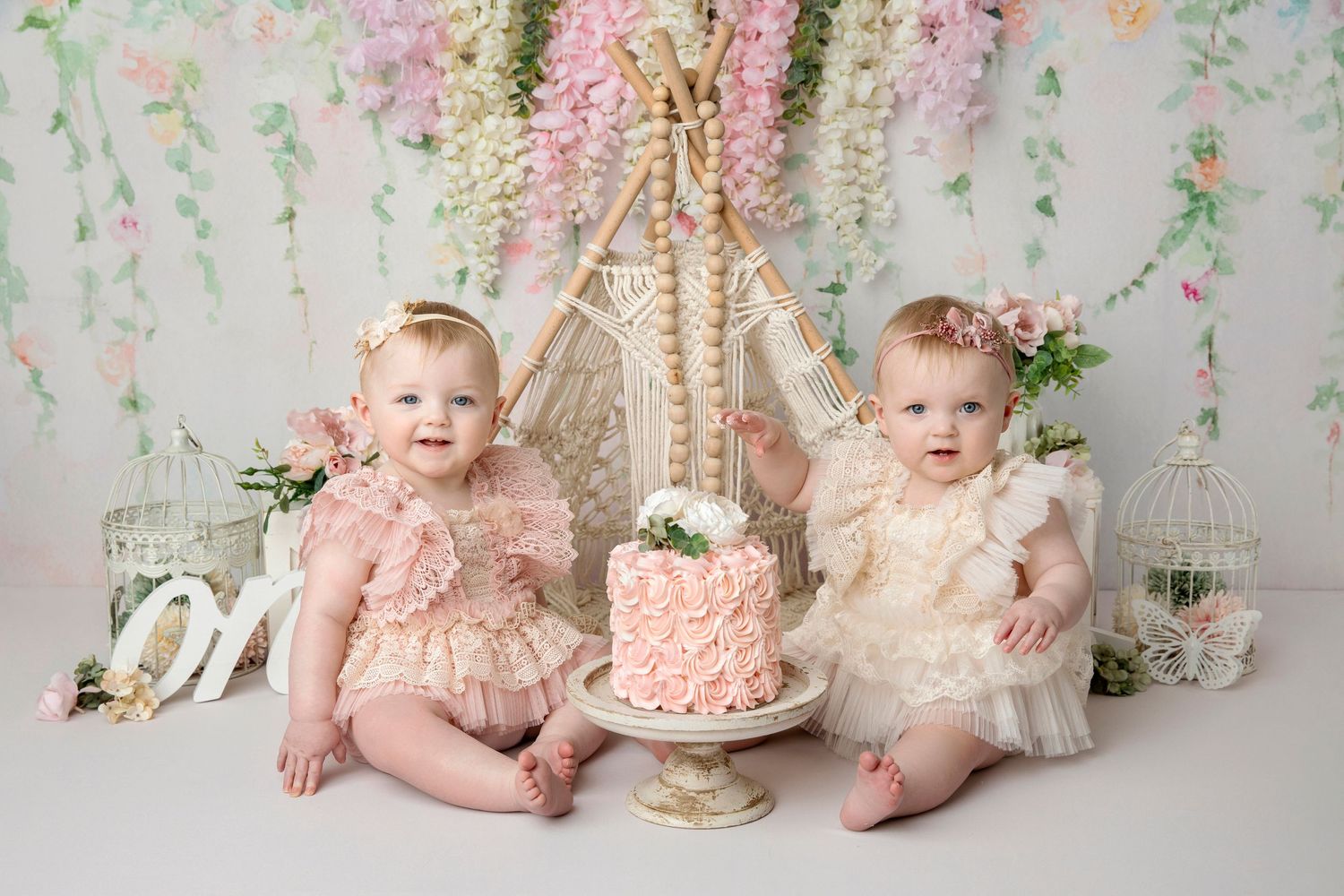 Two babies in vintage lace dresses seated with pink cake for first birthday photo session.