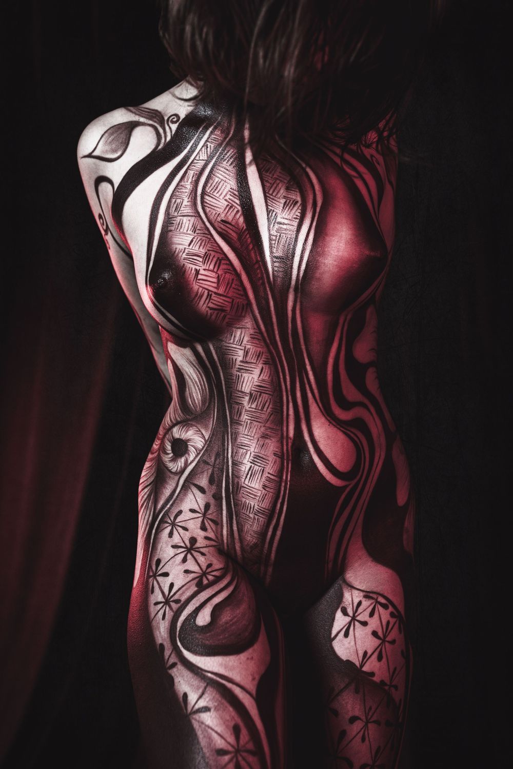 Intricate red and black body paint artwork covering female back and legs in dark studio.