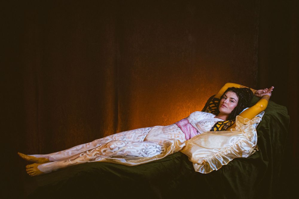 A woman in a white lace gown reclines gracefully on a velvet chaise lounge in dramatic warm lighting.