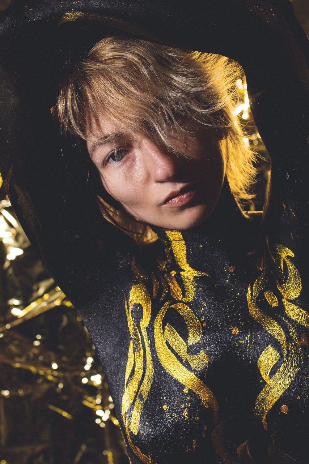 Woman with blonde hair wearing black and gold body paint poses dramatically against a dark golden background.