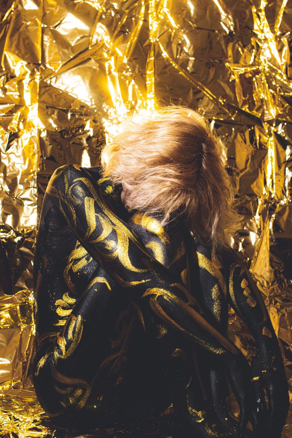 Person in black and gold outfit sitting against shimmering gold foil background with blonde hair.