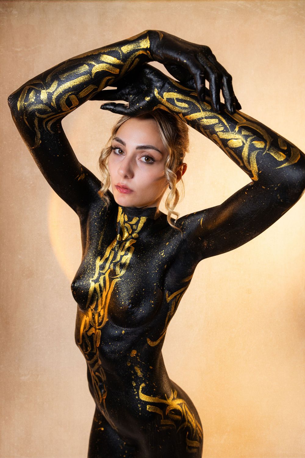 Woman covered in black and gold body paint with intricate patterns posing against a warm beige background.