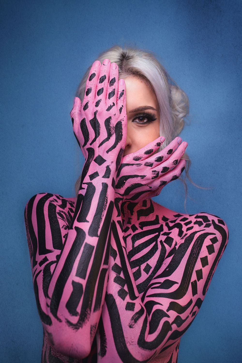 Woman with silver hair covered in pink and black geometric body paint poses on blue background.
