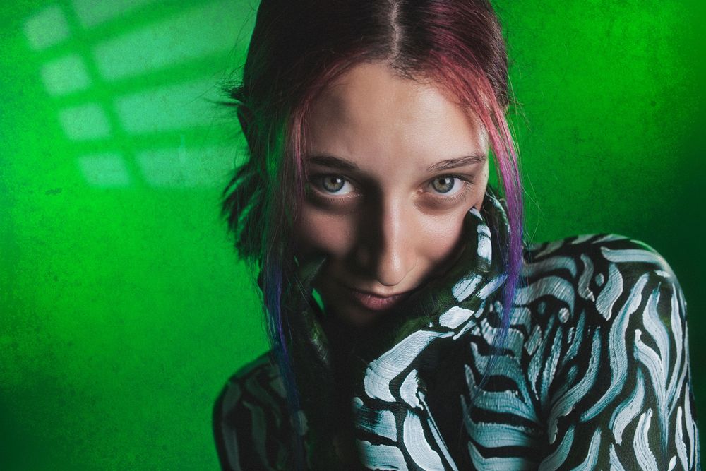 Young woman with colorful hair in zebra print bodysuit poses against vibrant green background.