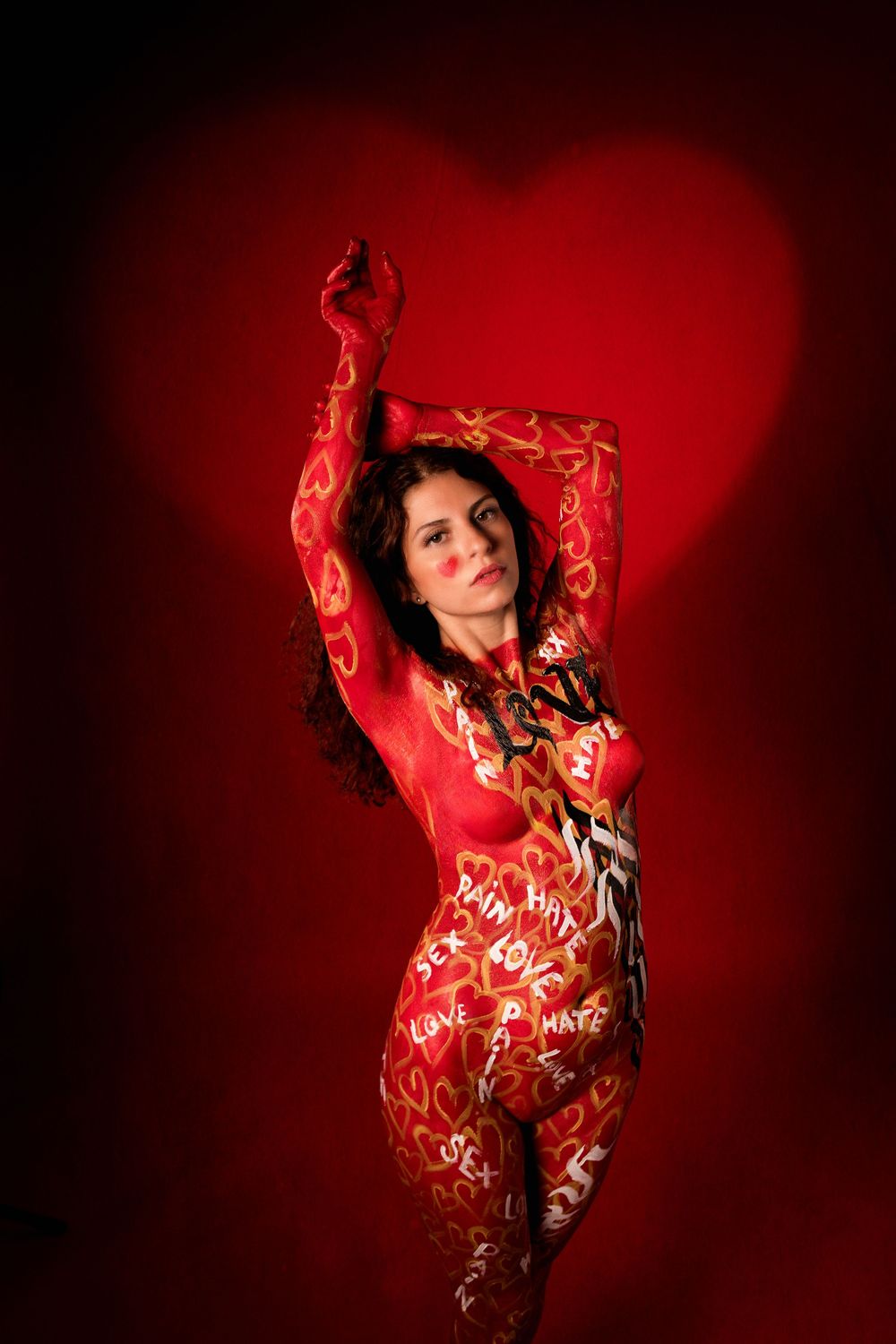 Female model with red and gold heart-themed body paint poses before glowing red heart backdrop.