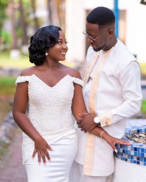 Couple in white formal attire pose together against outdoor setting.
