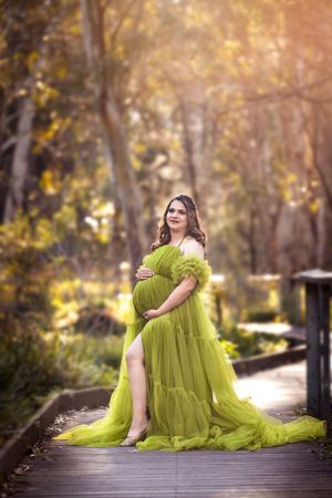 Maternity portrait of woman in lime green flowing dress on wooden bridge in autumn forest.