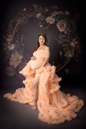 Elegant maternity photo with a floral arch backdrop and flowing peach-colored tulle gown.