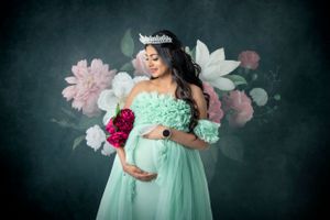 Maternity portrait with floral backdrop showing woman in mint green dress holding bouquet.