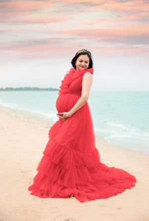 Maternity beach portrait with woman in flowing red dress against sunset sky.