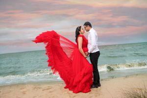 Romantic beach photoshoot with flowing red dress billowing in the wind against sunset sky.